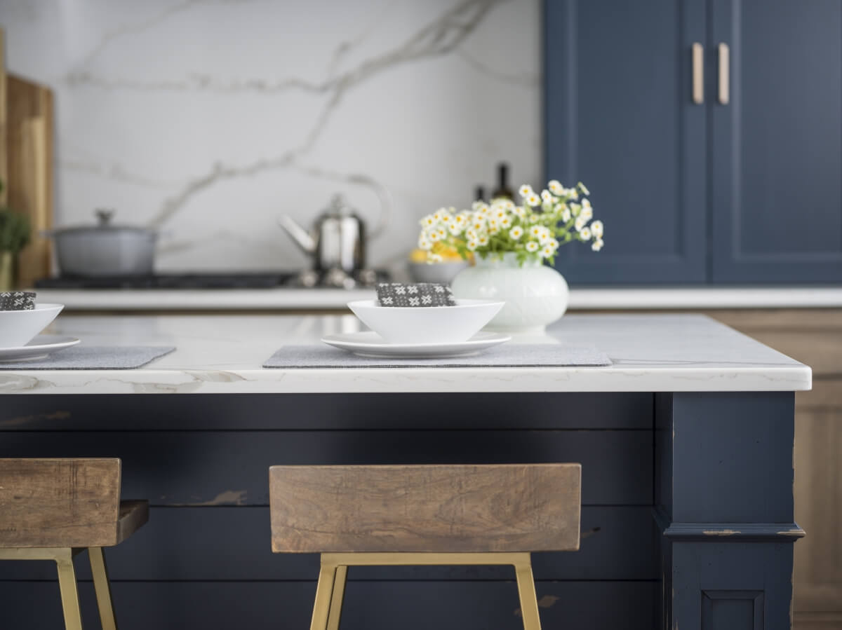 A close up of a navy blue kitchen island by Dura Supreme with gold bar stools and countertop height seating in the kitchen.
