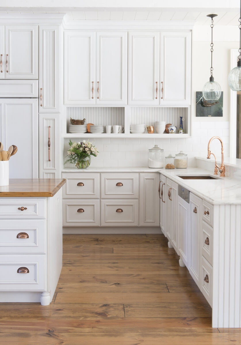 A bright white french farmhouse style kitchen with gold and brass hardware, white painted cabinets, white subway-tile backsplash and light stained wood floors.