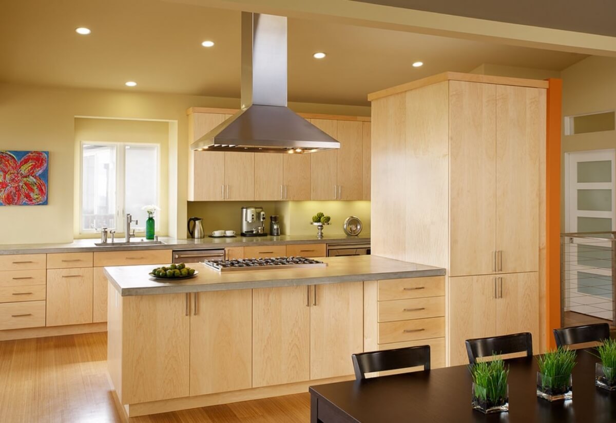 A modern styled kitchen with warm, light stained wood cabinets and a long kitchen peninsula with a cooktop and floating metal hood.