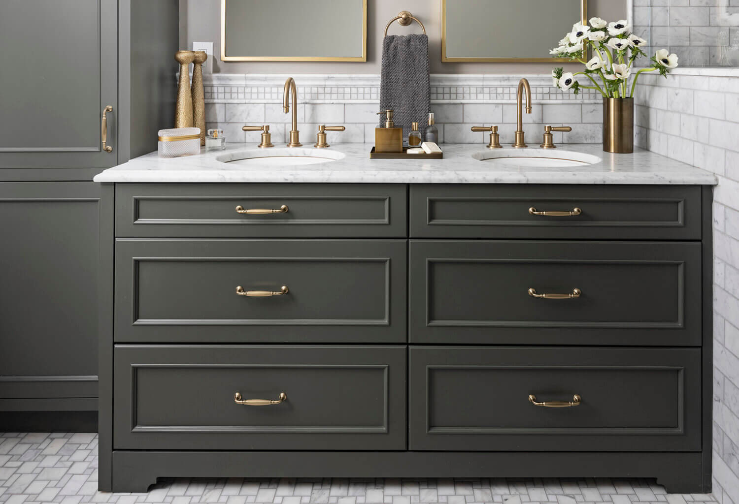 A dark green (almost black) painted bathroom vanity and tall linen cabinet with brass hardware and light gray tiles.