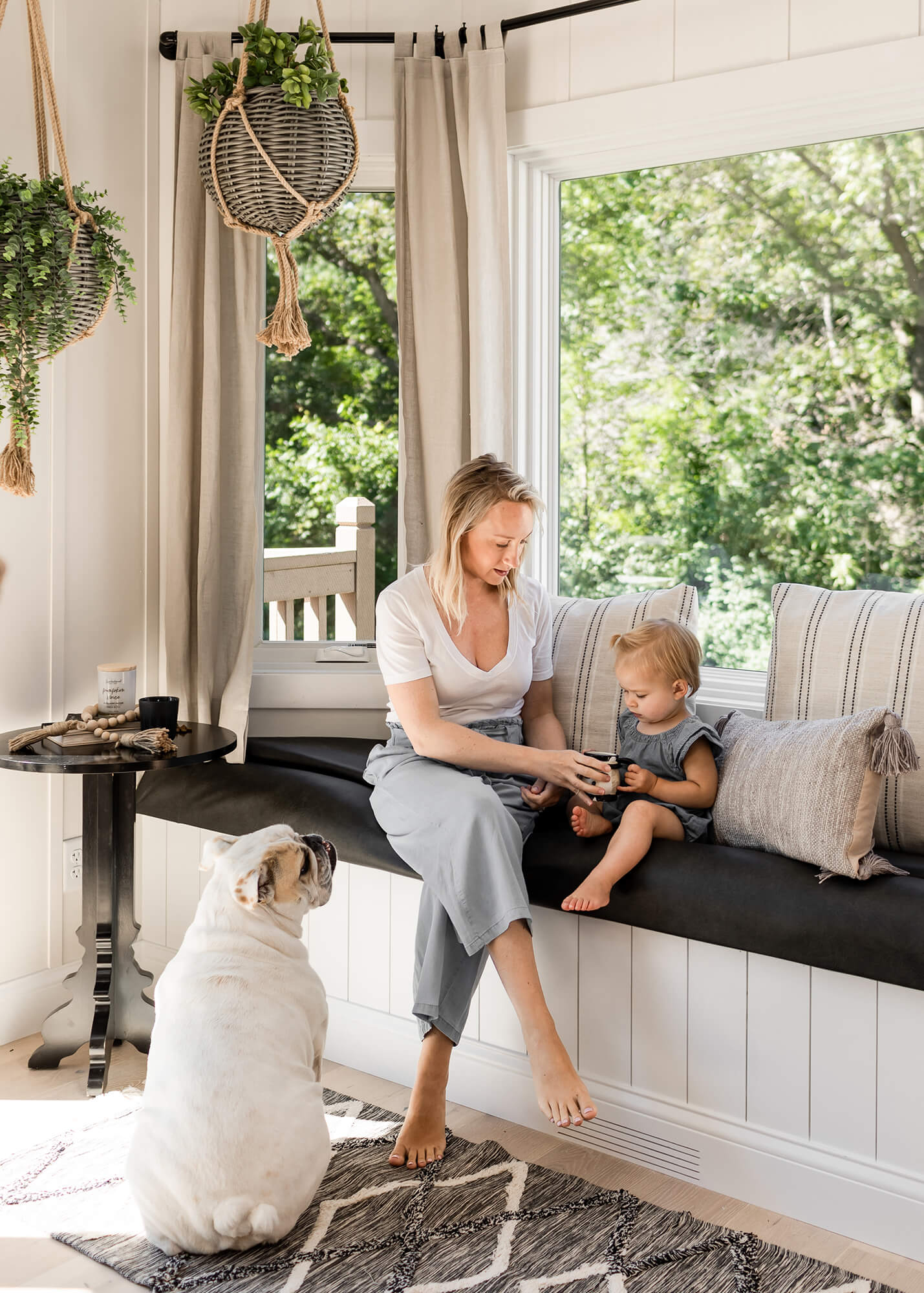 The principals behind Biophilic interior design are focused on crafting an environment that actively supports your wellness and reconnects you with the natural rhythms that ground us all. Mother, child, and dog sit in a window seat surrounded by natural light and greenery.