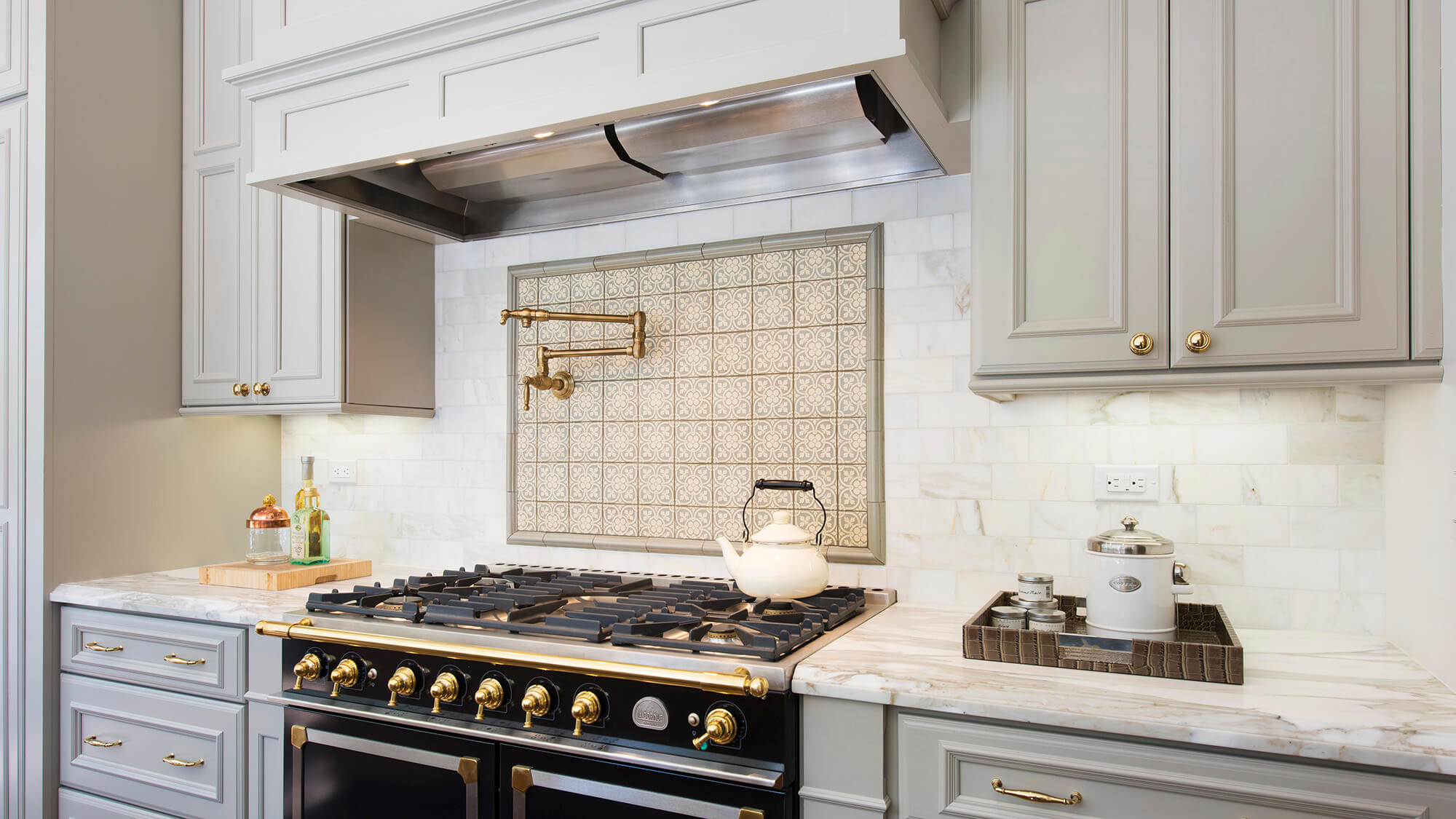 Beautiful slide-in, black and gold range with double ovens showing gray painted cabinets paired with brass cabinet hardware and fixtures. Featuring Dura Supreme kitchen cabinets and traditional styled wood hood canopy.