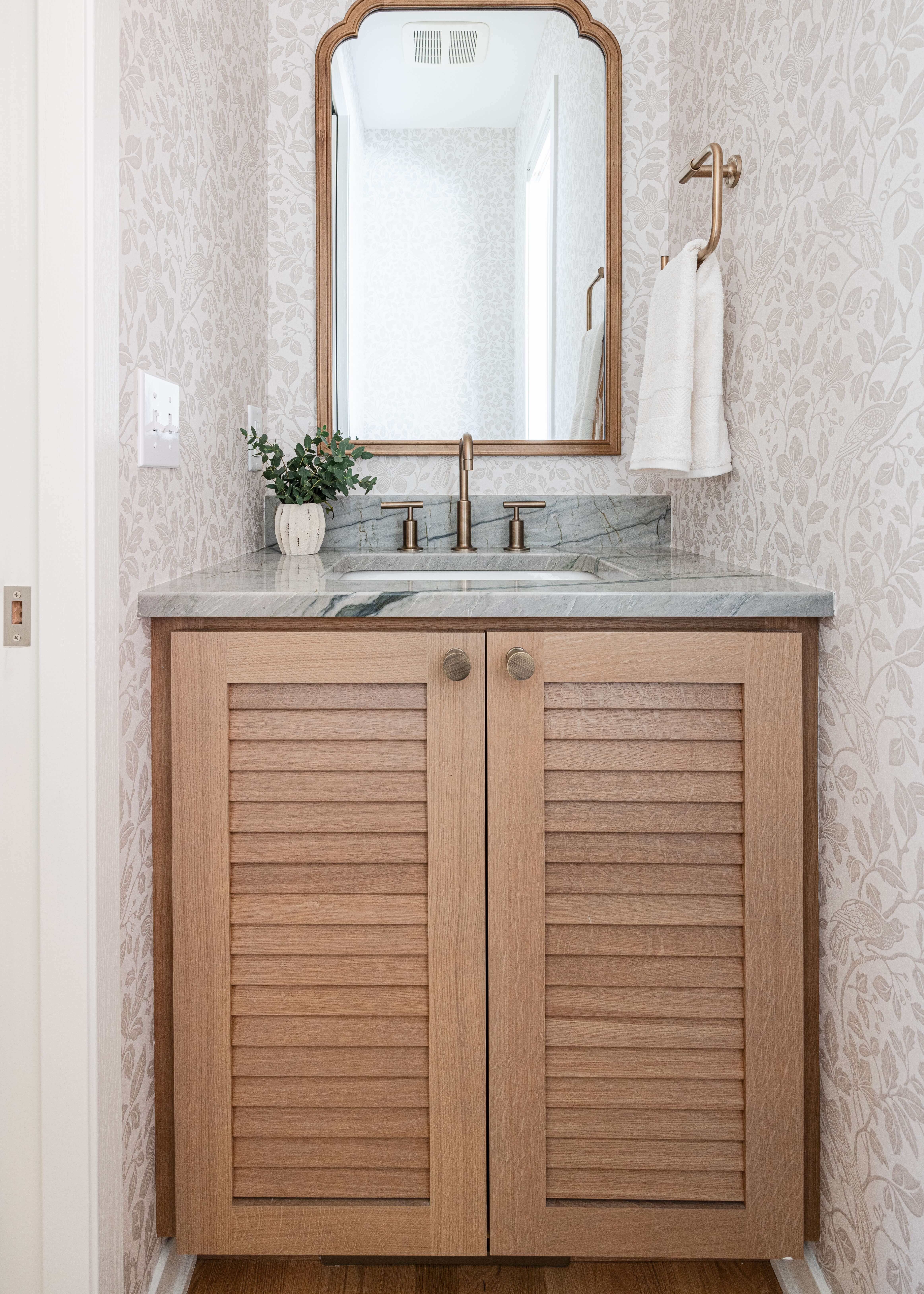 Light stained quarter-sawn white oak bathroom vanity cabinets showcase louvered cabinet door detailing and elegant round cabinet knobs. Natural wood tones, a stone countertop, and furniture-inspired styling bring warmth, texture, and timeless appeal to the bathroom.