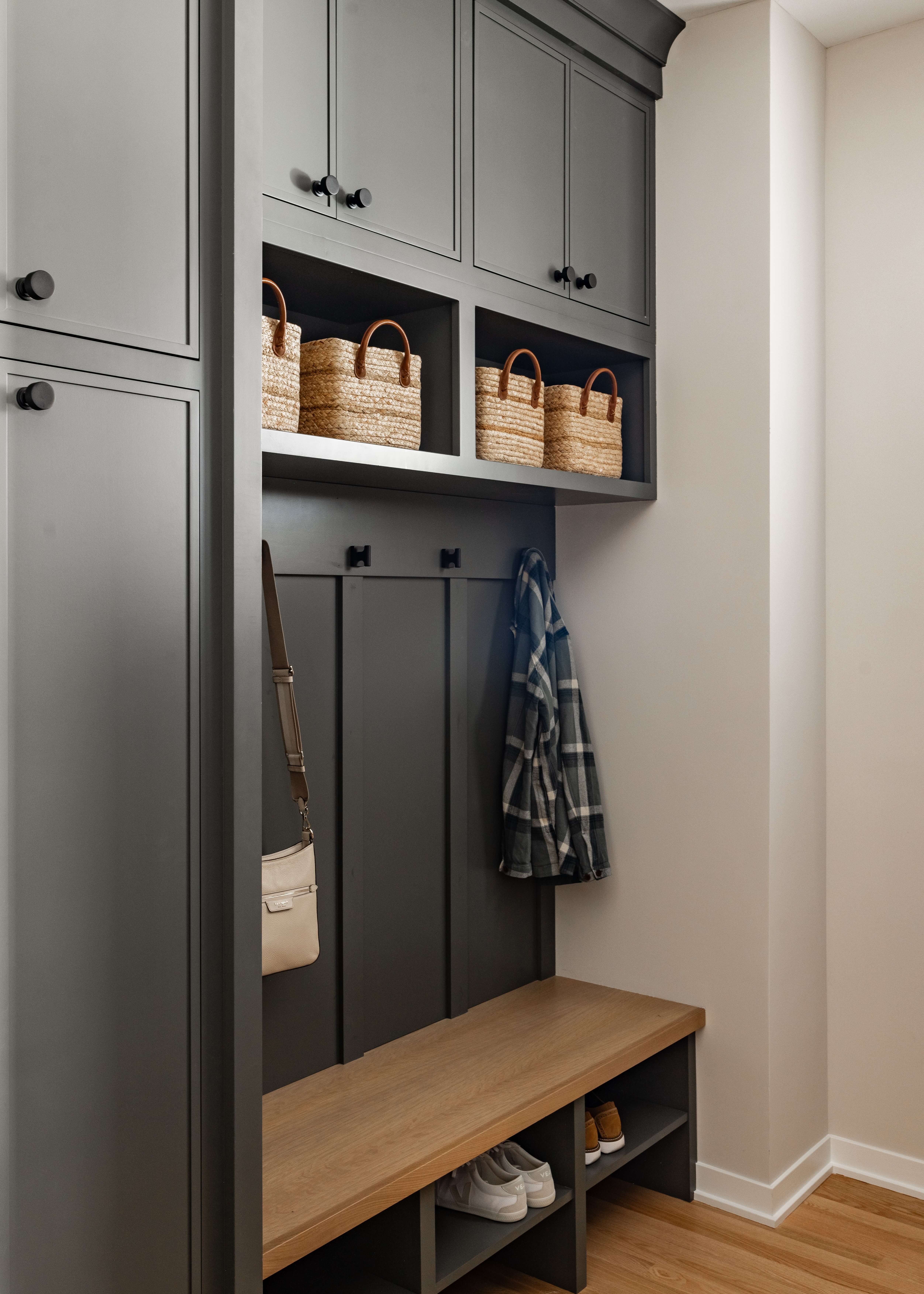 Mudroom lockers and a boot bench in a dark green painted finish create a stylish, organized entryway. Open cubbies, upper storage cabinets, coat hooks, and black cabinet hardware provide dedicated storage for shoes, outerwear, bags, and everyday essentials.