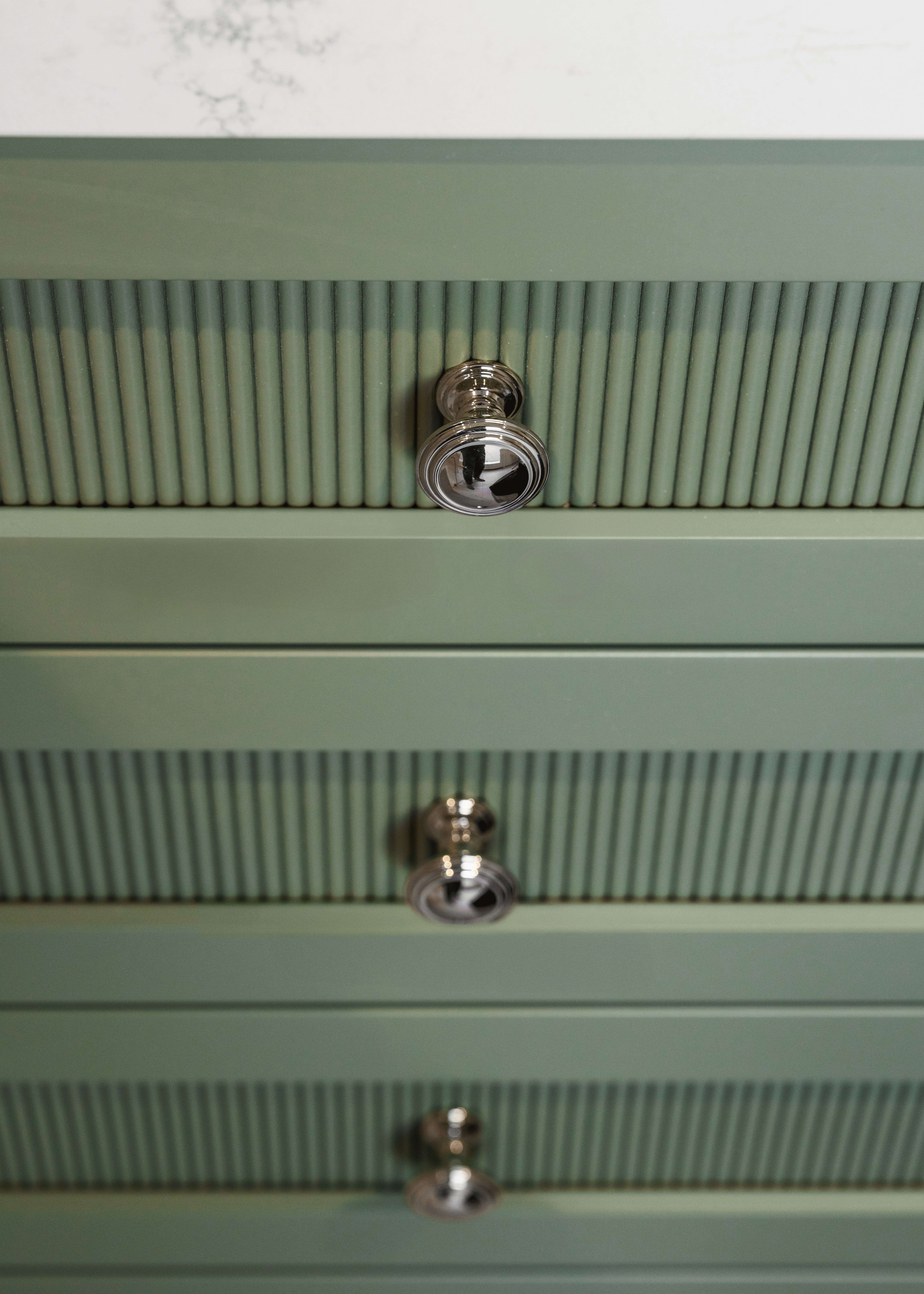 Light green painted custom kitchen cabinets with reeded cabinet door panels create rich texture and visual interest. Decorative round cabinet knobs add a classic touch, complementing the trend-forward color and detailed cabinetry design.