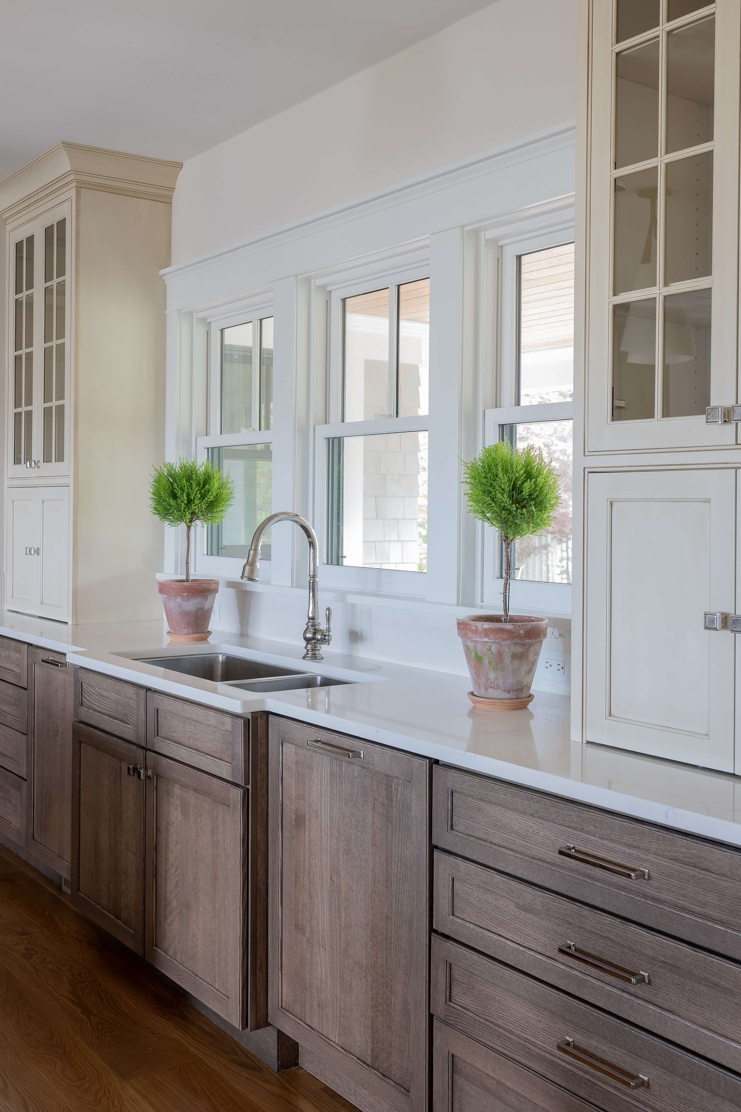 Two-tone custom kitchen cabinets blend rich stained wood base cabinets with bright white painted upper cabinets for a warm, timeless design. A thoughtfully curated mix of brass cabinet knobs, pulls, and latches adds character, while glass-front cabinets provide elegant display storage.