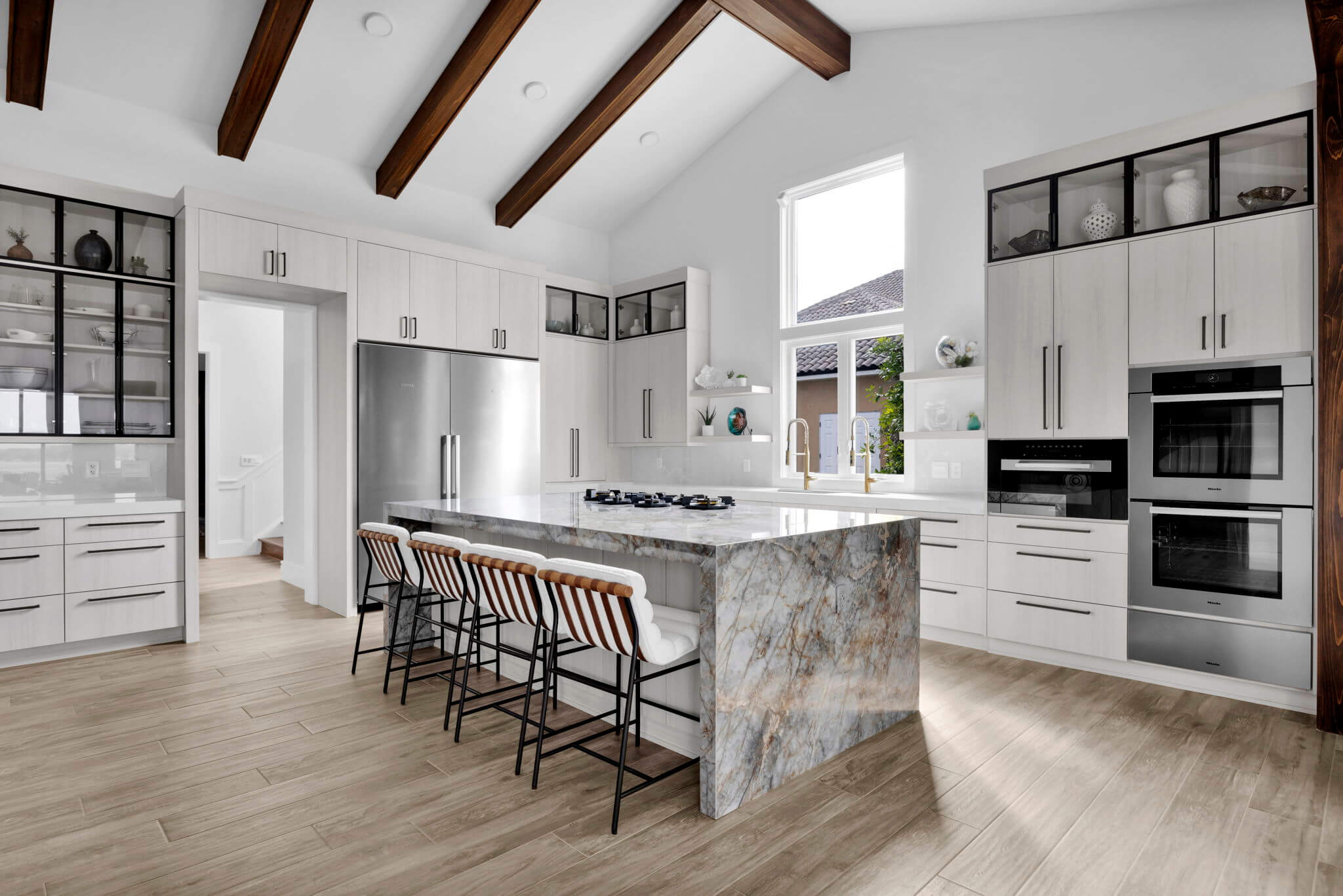 Premium kitchen cabinets in a bright white finish create an airy, open kitchen beneath dramatic vaulted ceilings with exposed wood beams. Sleek modern cabinet hardware, glass-front display cabinets, and a large kitchen island add timeless style and functionality.